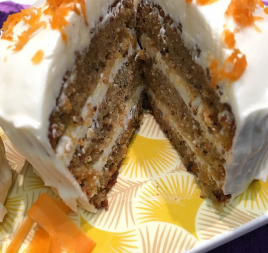 Triple layer gluten free carrot cake with a light fluffy frosting topped with fresh carrot shavings on a yellow and white plate.