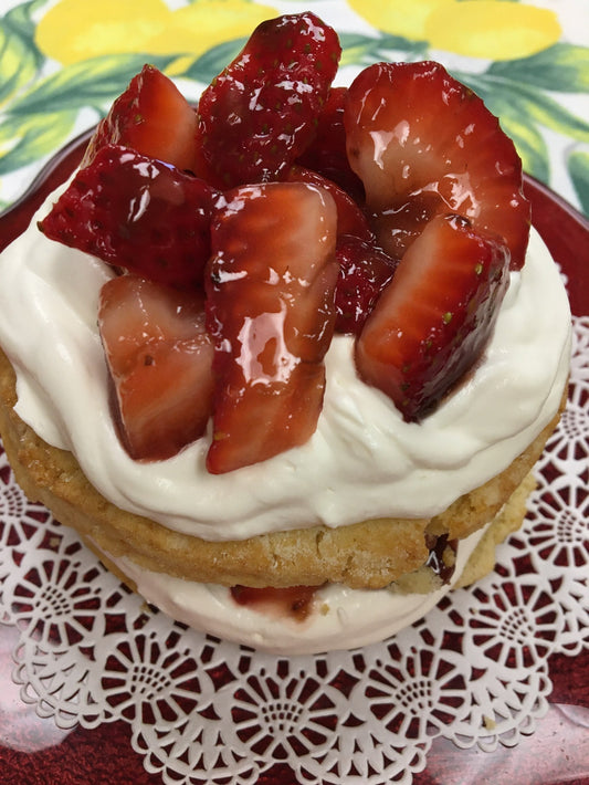 shortcake topped with fresh whip cream and fresh strawberries on a doily covered plate.