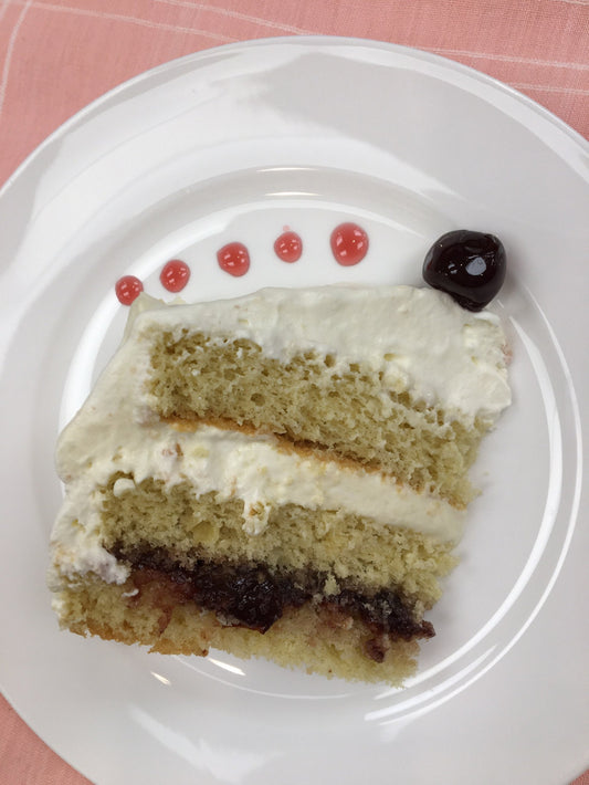 Three layers of sponge cake filled with fruit filling and cream. Topped with a cherry on a white place with pink tablecloth background.