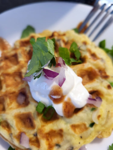 Golden gluten free waffle on a white plate topped with sour cream, red onions, cilantro and a few dollops of hot sauce.