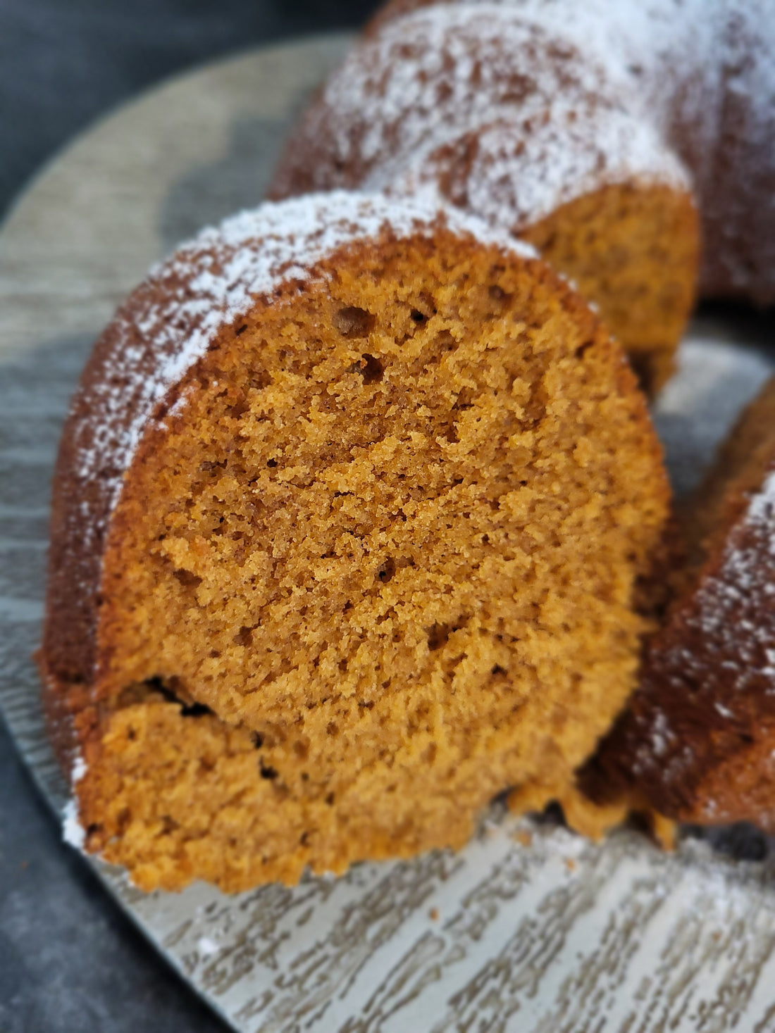A golden slice of gluten free gingerbread bundt bake topped with dusting of icing sugar on a grey wooden plate.
