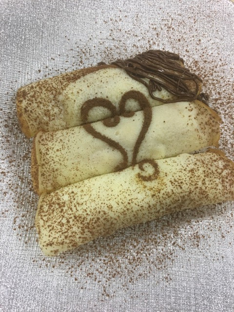 Three crepes rolled on white and silver plate with a heart design made with cocoa powder and the top right edge drizzled with chocolate.
