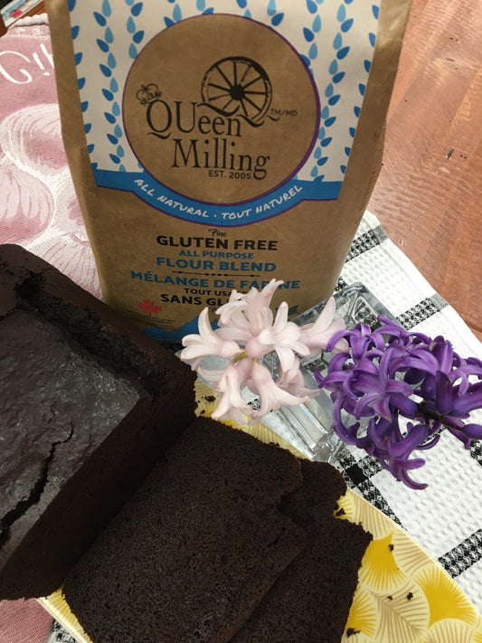 gluten free dark chocolate loaf sliced on a yellow plate.  purple and pink flowers on table and a QUeen Milling All Purpose Flour Bag standing beside gluten free loaf.