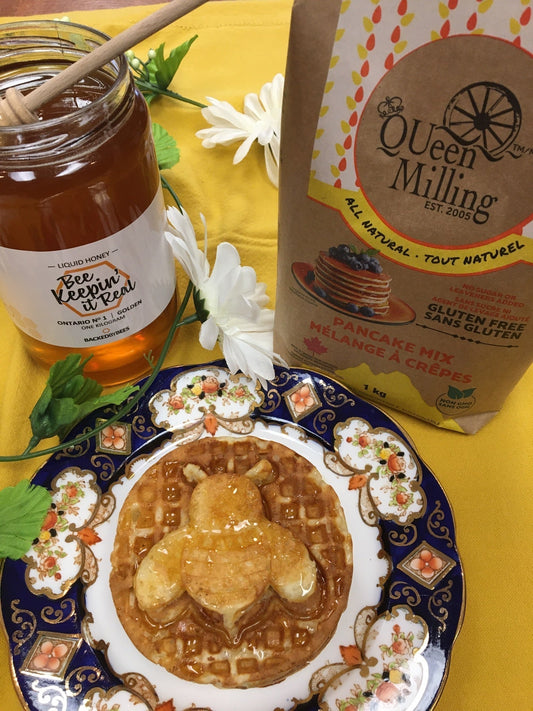 a bumblebee character waffle baked golden and topped with Ontario honey.  All on a blue colour plate.  Honey jar open beside a bag of QUeen Milling Superfine Gluten Free Pancake Mix.