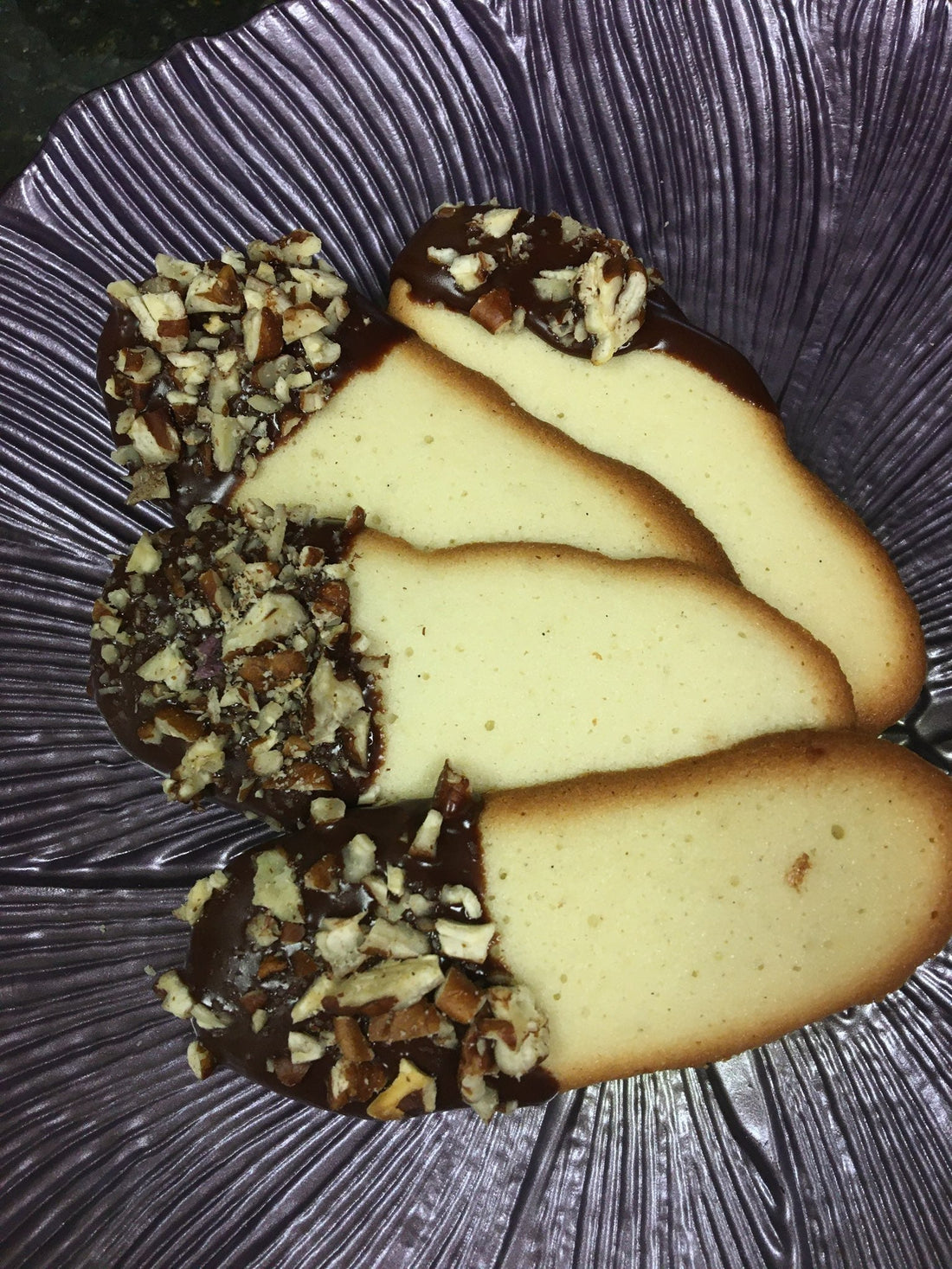 Gluten free langues de chat cookies baked golden brown and dipped in a chocolate ganache.  Sprinkled with chopped nuts of your choice.  Four cookies on a purple spring petal plate.