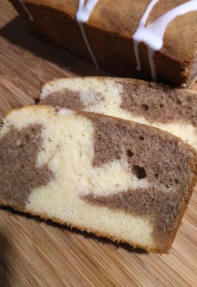 A white and brown swirl piece of cake on a wooden board.  Loaf in background drizzled with icing sugar glaze.