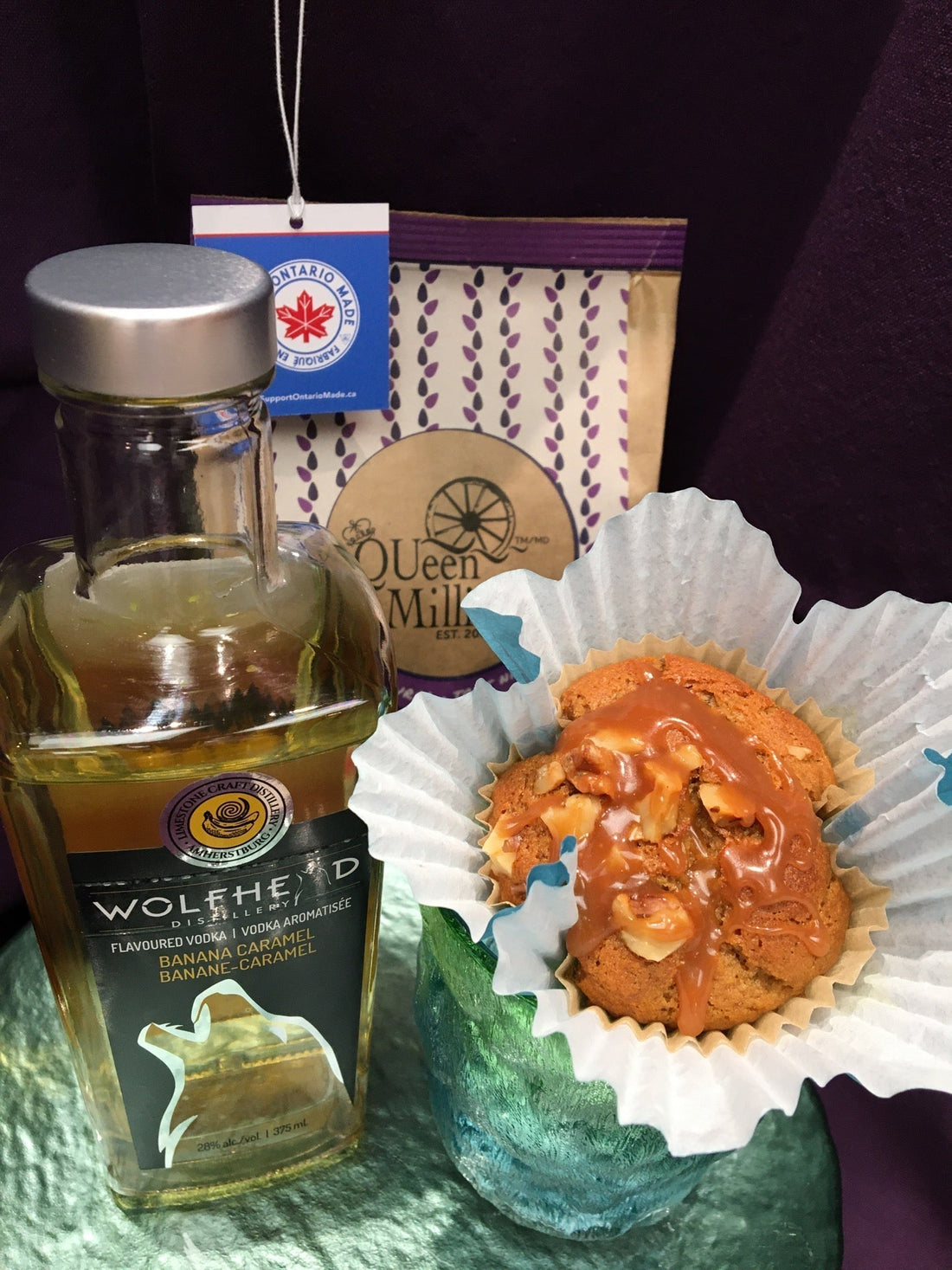 Carmel topped muffin beside a bottle of Wolfhead Banana Caramel Vodka and a bag of QUeen Milling Gluten Free Wholegrain Brown Rice Flour behind. The bag is kraft paper with purple and an Ontario Made tag hanging in the corner.