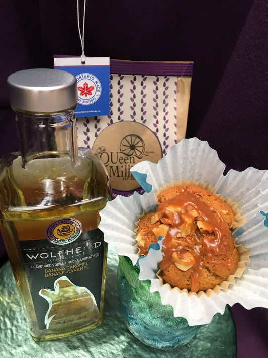 Carmel topped muffin beside a bottle of Wolfhead Banana Caramel Vodka and a bag of QUeen Milling Gluten Free Wholegrain Brown Rice Flour behind. The bag is kraft paper with purple and an Ontario Made tag hanging in the corner.