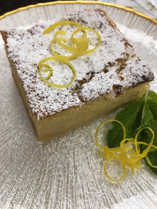 Square piece of cake on top of a silver white plate.  Topped off with icing sugar and lemon zest and green mint for accent.