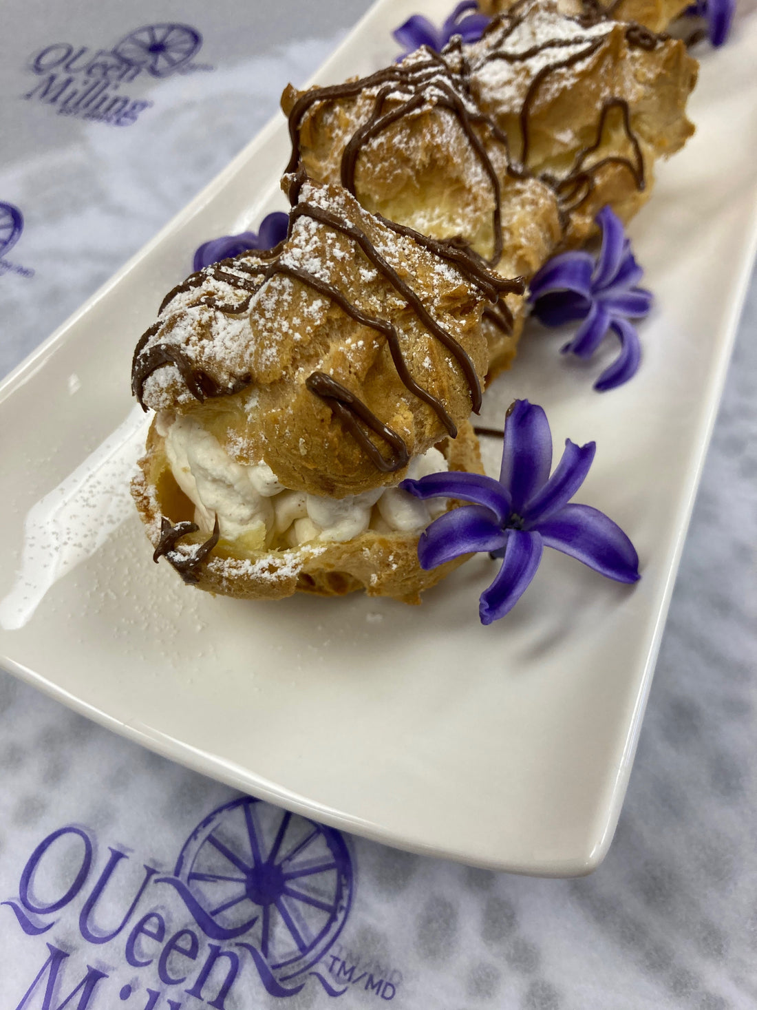 Round cream puffs on a white place which is placed on top of QUeen Milling logoed paper.  Cream puffs are drizzled with chocolate and purple flours line the plate.
