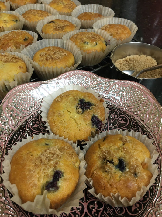 golden gluten free blueberry muffins on a pink plate with a dozen gluten free muffins pictured behind.  Baked with QUeen Milling Gluten Free All Purpose Flour Blend.