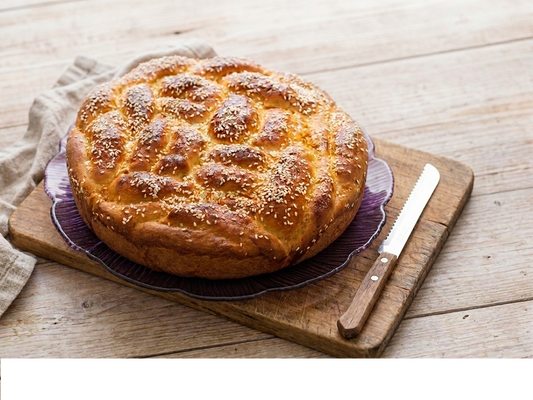 Round gluten free braided challah loaf on a wooden cutting board, napkin on the left, bread knife placed on the right of the loaf.