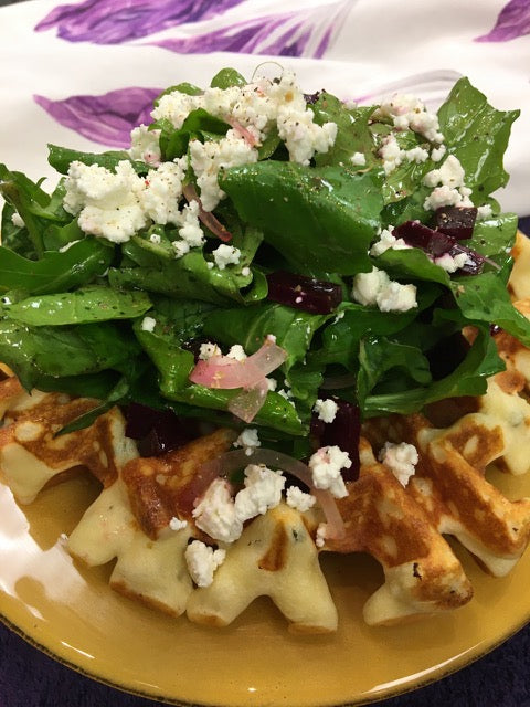 Large waffle on a gold plate, topped with fresh salad and goat cheese.
