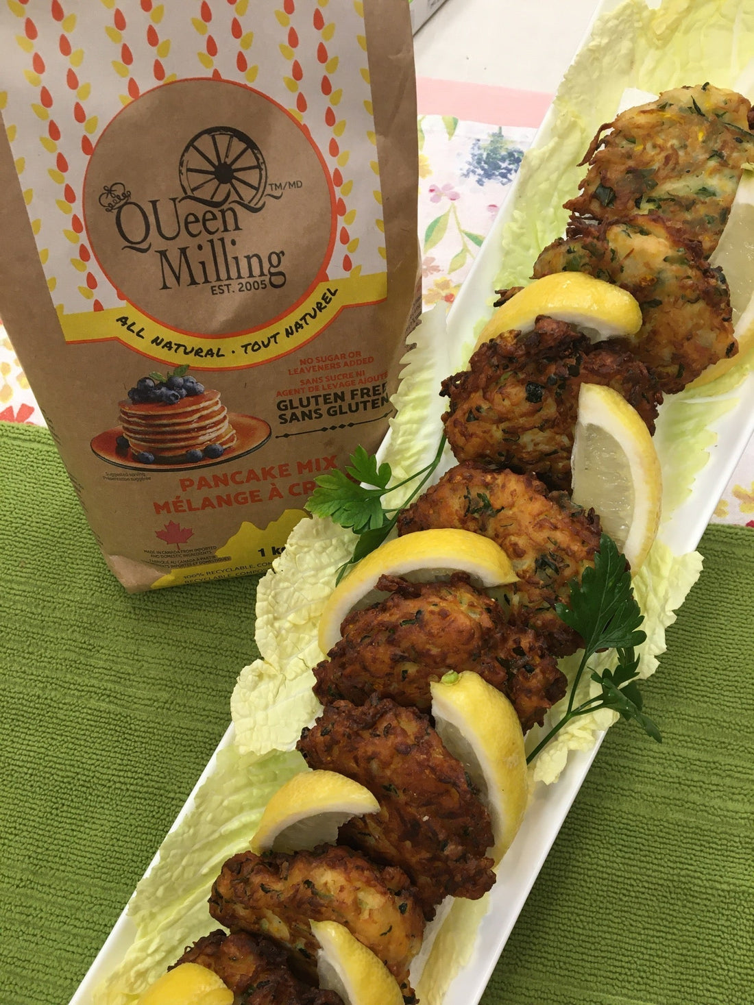 Gluten free zucchini fritters lined on an oblong white plate with lemon and parsley.  A bag of QUeen Milling Made in Ontario Gluten Free Pancake Mix beside it.  All on a green cloth.