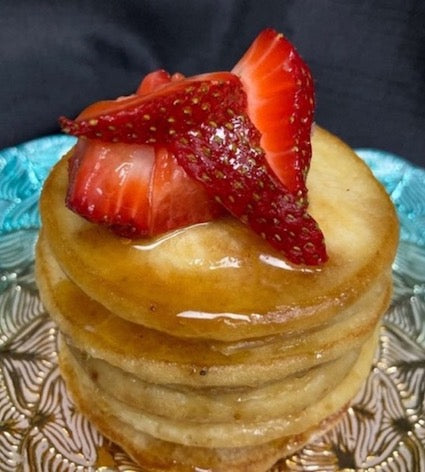 5 mini pancakes stacked on a colourful white and blue plate.  Pancakes topped with fresh ripe red strawberries and drizzled with syrup.