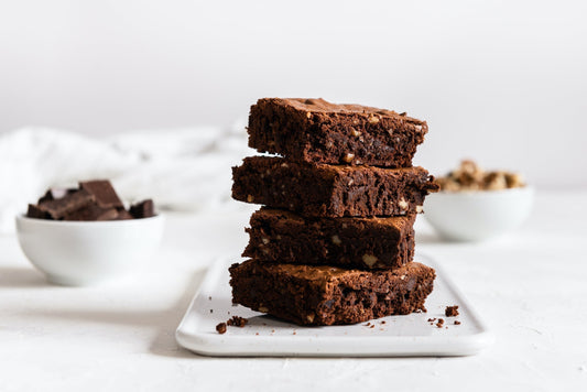 Four brownie squares stacked on a white plate.  A bowl with chocolate and one with nuts in the background.
